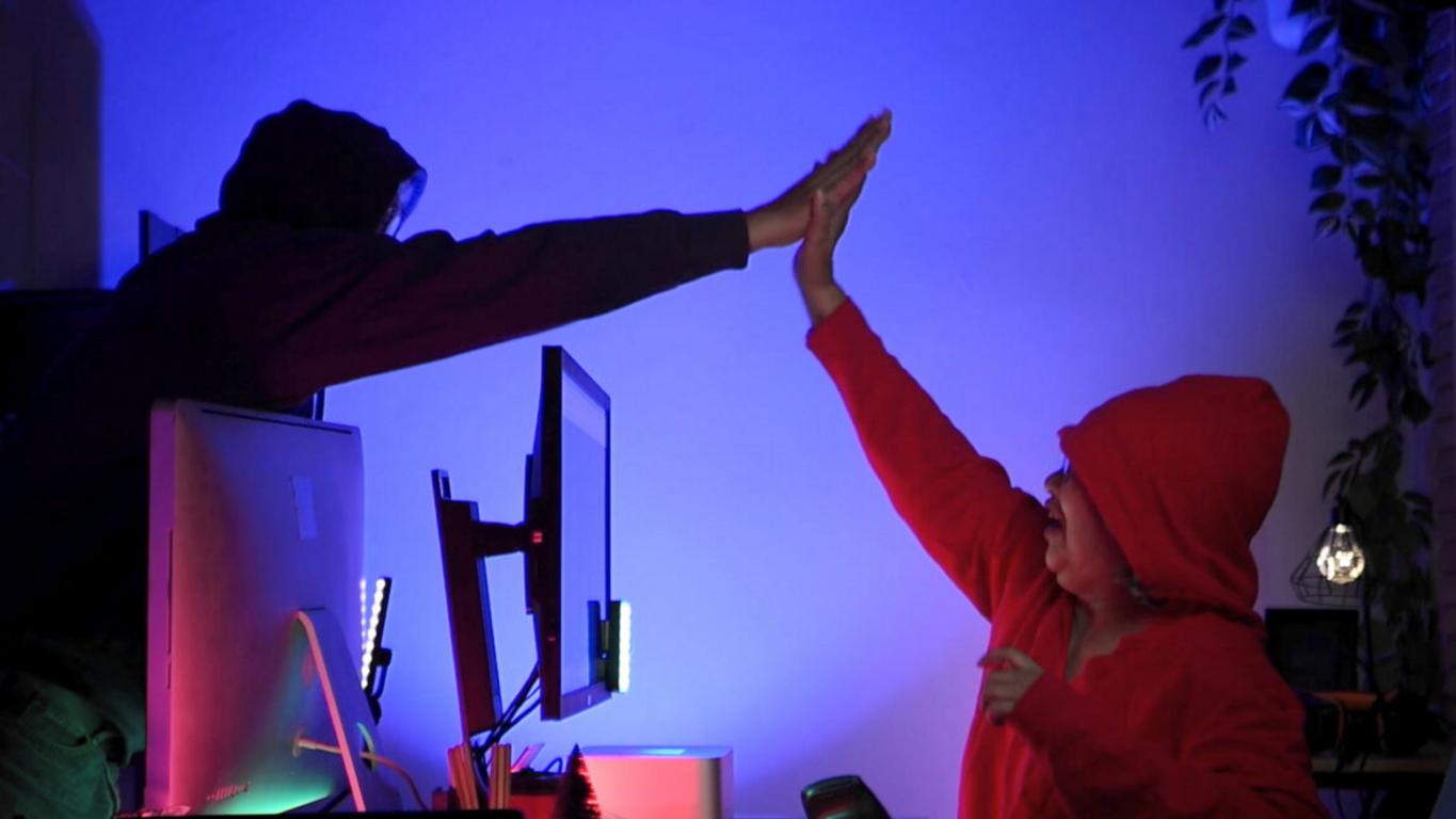 Dos supuestos hackers chocando los cinco frente al ordenador en una sala iluminada con luces de neón