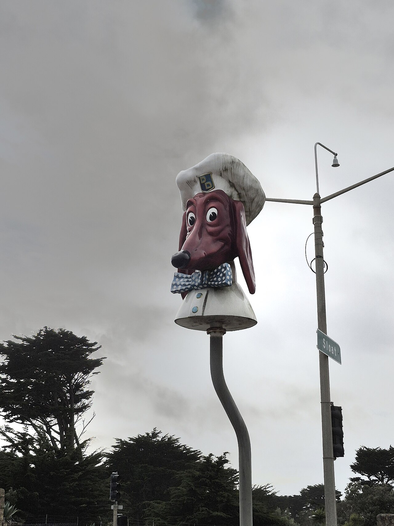 Cabeza de perro salchicha con gorro de cocinero y pajarita de lunares sobre un poste curvo, bajo un cielo gris