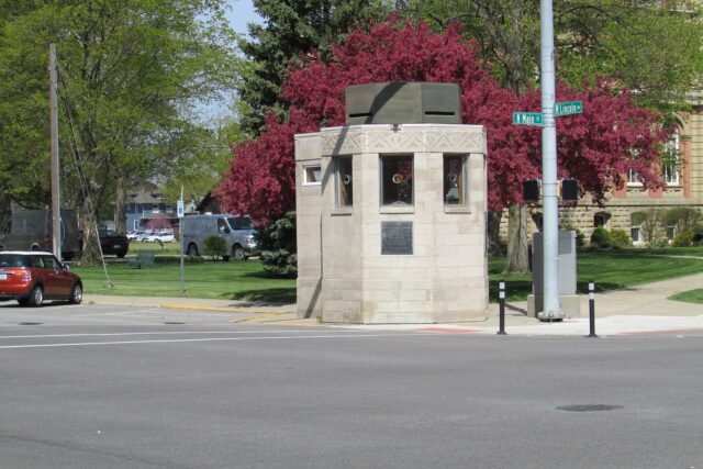 Caseta antibalas de la policía de Goshen, Indiana Caseta octogonal de piedra clara con grandes ventanales en una esquina de calle, junto a los rótulos N Main St y W Lincoln Av