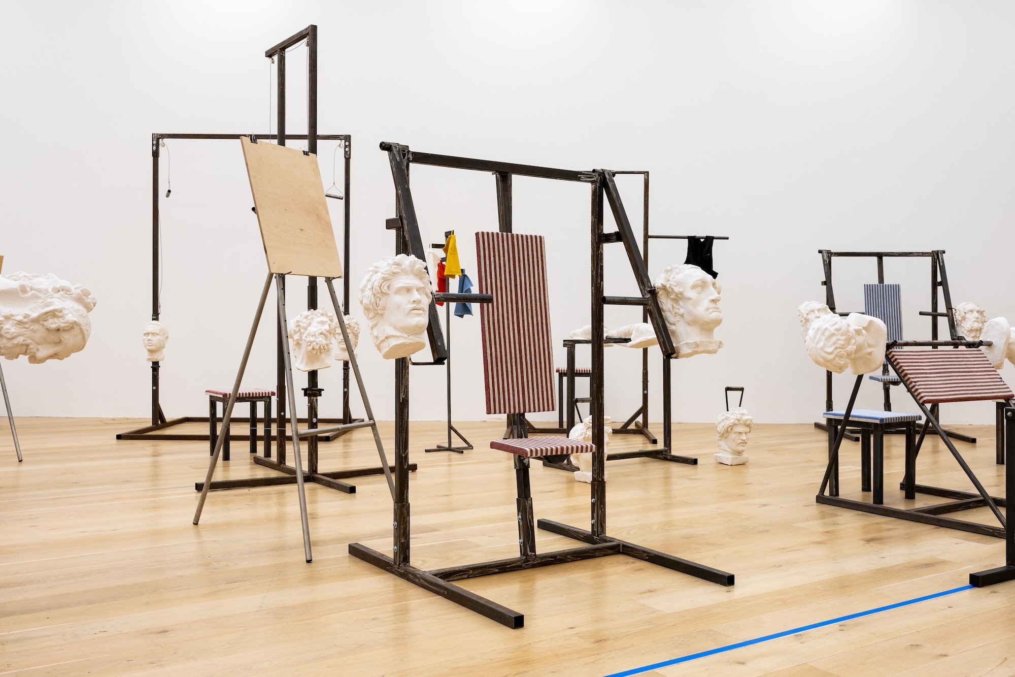 Sala de museo con máquinas de gimnasio de metal negro cuyos discos y contrapesos son bustos clásicos blancos