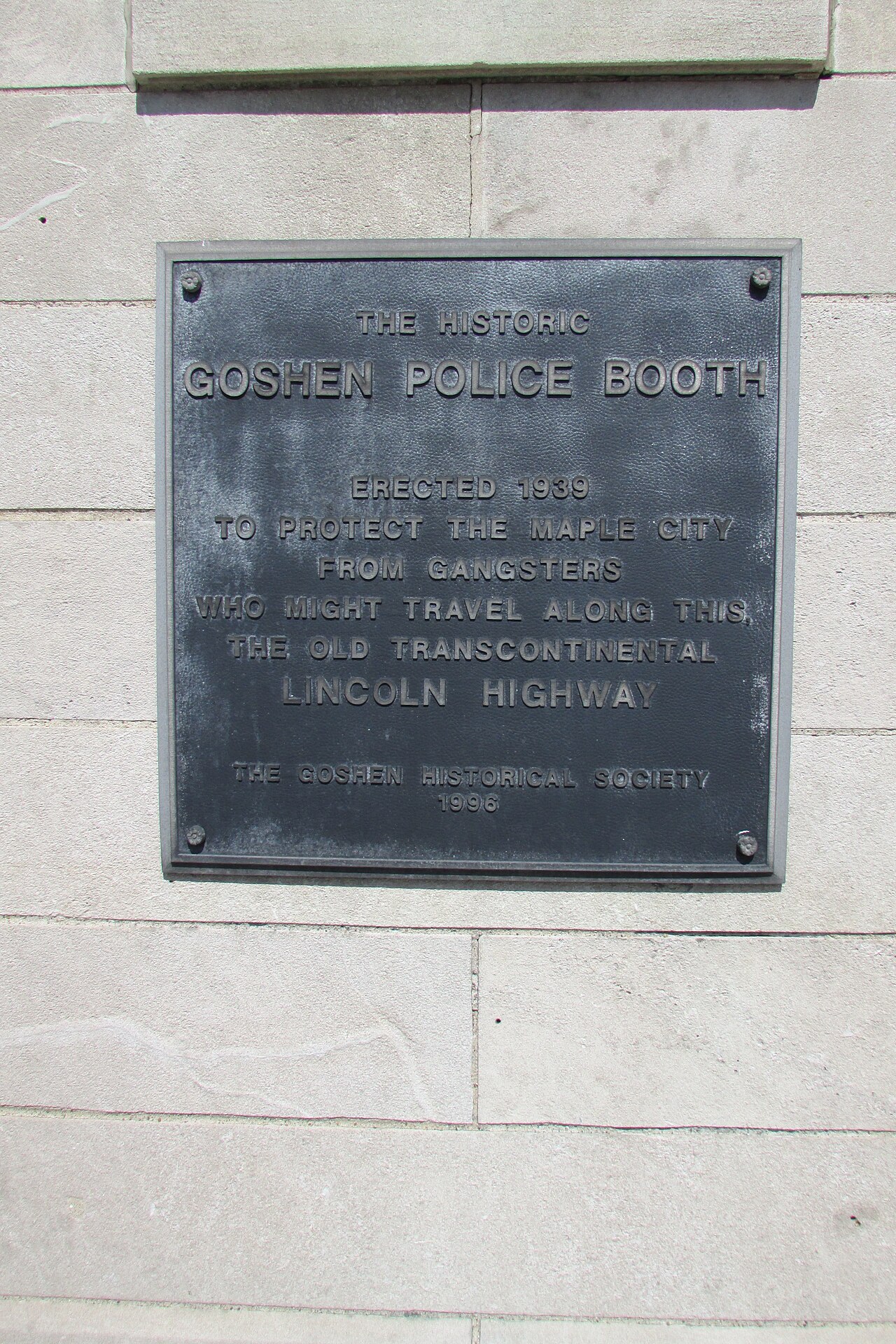 Placa metálica oscura atornillada al muro de piedra de la caseta con la inscripción The Historic Goshen Police Booth, erected 1939
