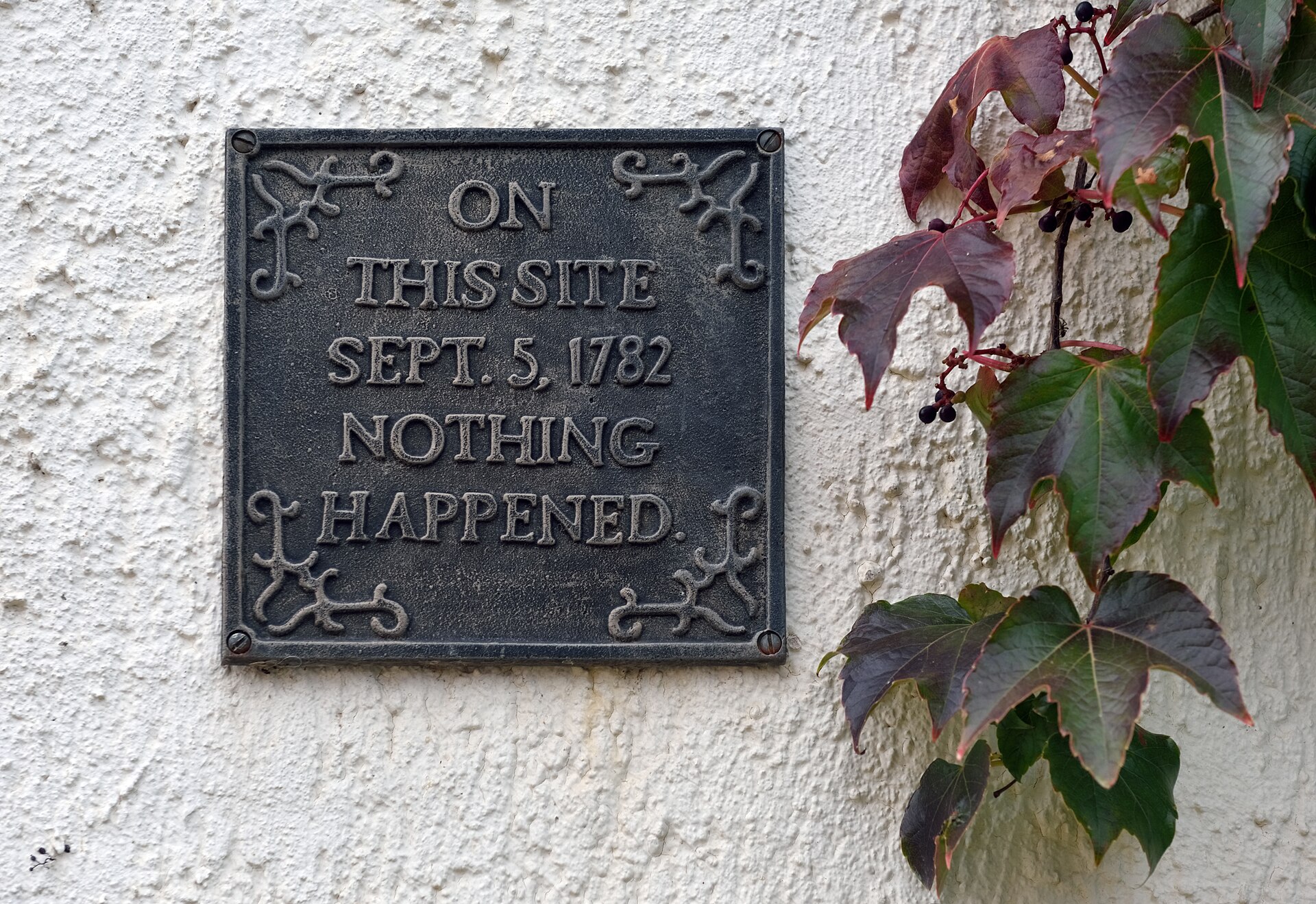 Placa de metal oscuro atornillada a una pared blanca junto a una enredadera, con el texto ON THIS SITE SEPT. 5, 1782 NOTHING HAPPENED
