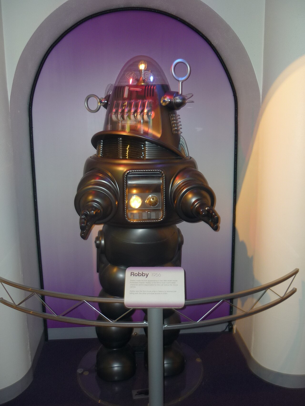 Réplica del robot Robby en una vitrina de museo, con cúpula de cristal, luces de colores y brazos articulados, junto a un cartel que pone Robby 1956