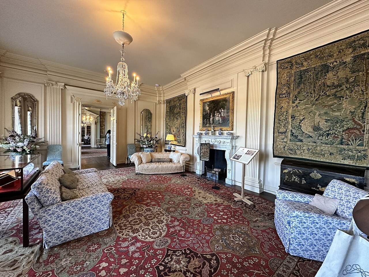 Salón señorial con sofás azules, alfombra roja, tapices, chimenea blanca y un atril informativo de museo junto a ella