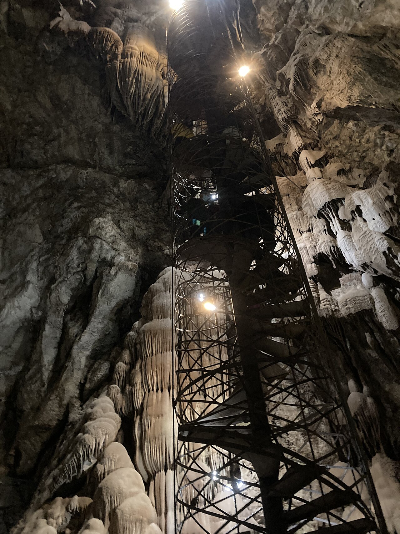 Escalera de caracol metálica iluminada que sube por la sala de Moaning Cavern entre coladas de calcita y estalactitas