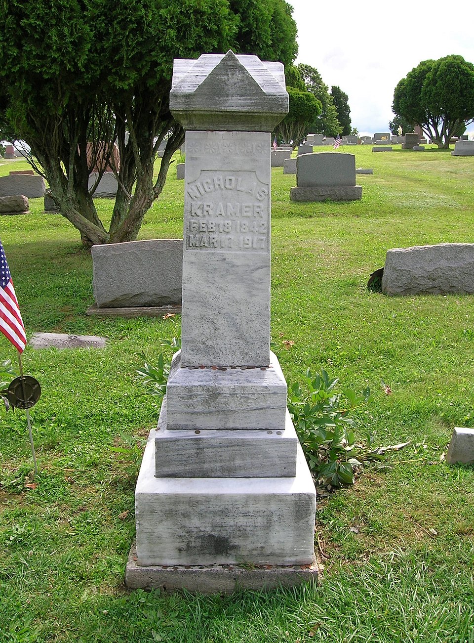 Estela funeraria de piedra clara con base escalonada, una pequeña bandera estadounidense clavada al lado y monedas sobre los escalones
