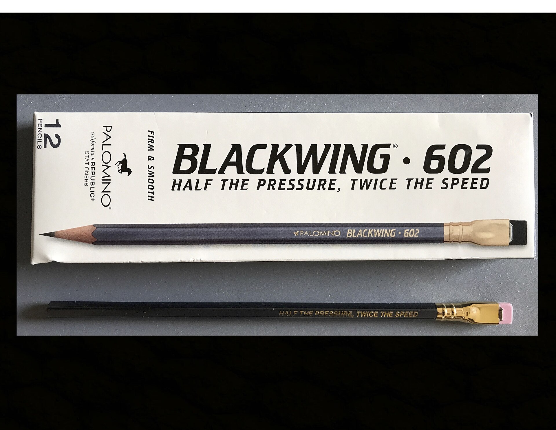 Caja blanca de doce lápices Palomino Blackwing 602 junto a dos lápices grises con virola dorada sobre fondo gris