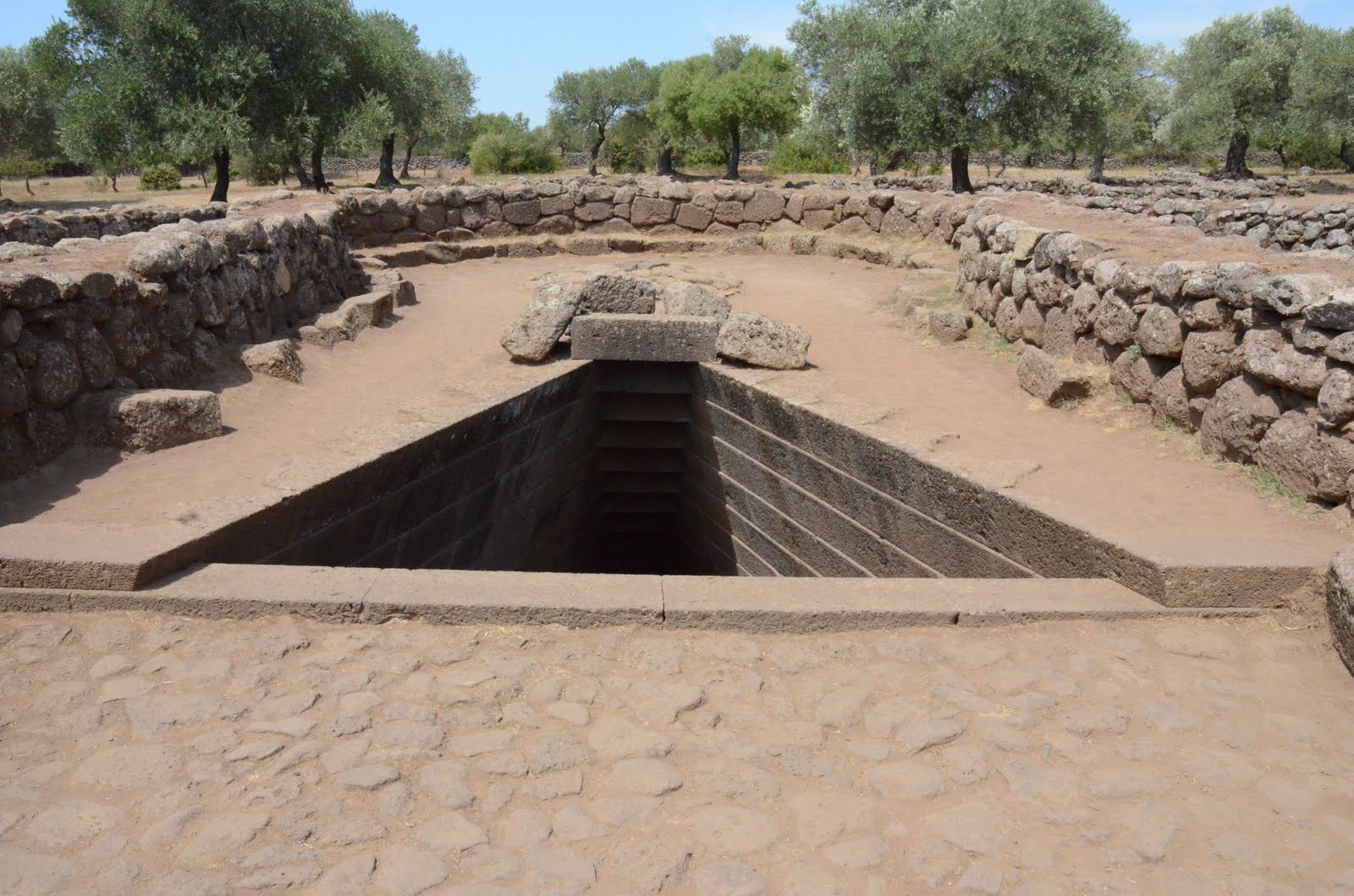 Escalera de piedra que desciende bajo tierra hacia el pozo sagrado de Santa Cristina, dentro de un recinto ovalado de muros entre olivos