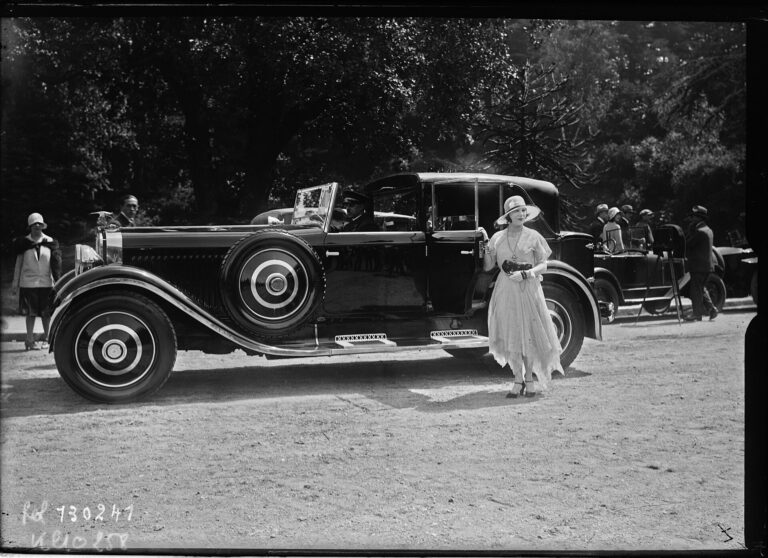 Mujer con vestido claro y sombrero cloche de pie junto a un gran automóvil Hispano-Suiza descapotable en un concurso de elegancia al aire libre en 1928