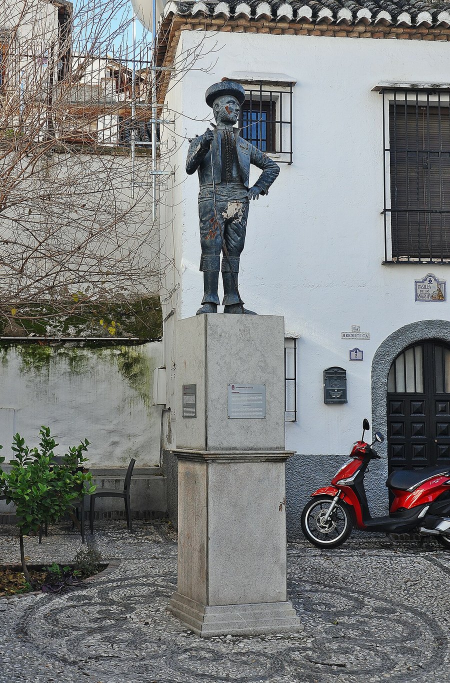Estatua de Chorrojumo con sombrero sobre un pedestal de piedra en una placeta empedrada de Granada, junto a una casa blanca